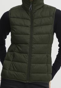 Vadderad väst i mörkgrön färg med hög krage och framdragkedja, med kviltade sektioner och en slät, lätt glänsande yta.