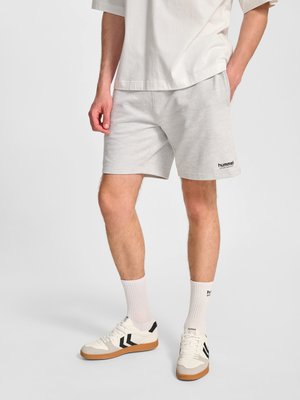 Man draagt lichtgrijze korte broek, witte sokken en witte sneakers met zwarte strepen, staand met handen in en bij de zakken.