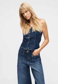Ξανθιά γυναίκα που φοράει χωρίς μανίκια τζιν τοπ με κουμπιά και τζιν παντελόνι φαρδιάς γραμμής, με το ένα χέρι στην τσέπη, ποζάροντας μπροστά σε λευκό φόντο.