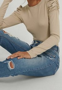 Beige gestreifter Langarmshirt mit gerafftem Schultern, kombiniert mit ausgewaschenen blauen Ripped Jeans, die verstörte Knieakzente und hohe Taille aufweisen.