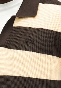 Gros plan sur un polo en maille avec de larges rayures horizontales marron et beige et un petit logo crocodile brodé sur la poitrine.