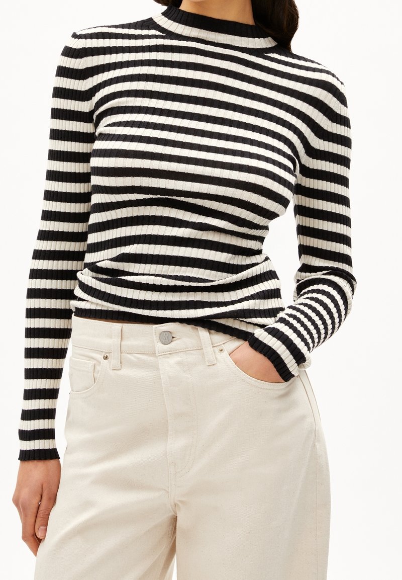 Femme portant un pull côtelé à rayures horizontales noires et blanches ajusté, avec un pantalon crème taille haute, une main dans la poche.