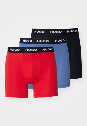 Trys HUGO trumpikių poros: raudonos, šviesiai mėlynos ir juodos, su juodu juosmeniu, ant kurio užrašytas baltas "HUGO" logotipas, pagamintos iš lygios medžiagos.