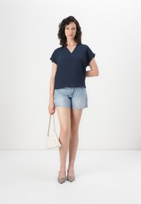 Navyblaues Kurzarmblusenshirt mit V-Ausschnitt, kombiniert mit hellen Denim-Shorts. Das Model hält eine kleine weiße Handtasche und trägt transparente Absatzschuhe.