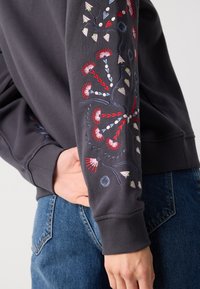 Felpa grigia scura con intricate ricamo floreale multicolore sul maniche sinistro e polsini a coste, abbinata a jeans di denim.