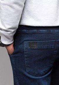 Pantalones de denim azul oscuro con un bolsillo trasero que presenta una etiqueta de cuero negro. La cinturilla es parcialmente visible, mostrando un detalle de costura en contraste.