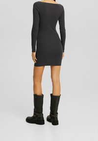 Femme debout de dos, portant une robe noire ajustée à manches longues et des bottes noires montantes avec des brides à boucles.