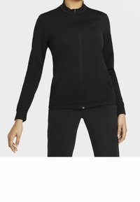 Kvinna i en figurnära svart zip-up jacka med långa ärmar och matchande svarta byxor, stående mot en enkel bakgrund.