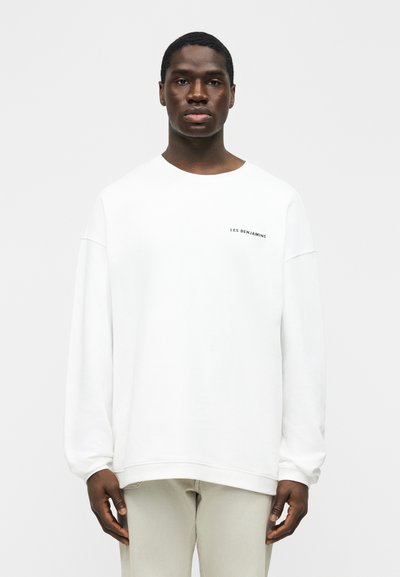 Νεαρός άνδρας που φοράει ένα απλό λευκό φούτερ με μακριά μανίκια και το κείμενο «LES BENJAMINS» στο στήθος, στέκεται μπροστά σε λευκό φόντο.