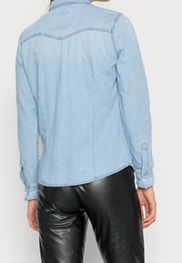 Ljusblå denimskjorta med spetsig krage, synlig sömnad och knäppte ärmar, kombinerad med svarta läderbyxor. Slät textur och rak passform.