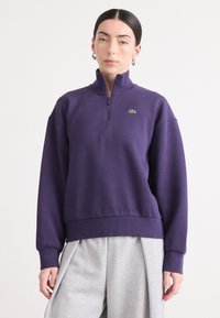 Paarse ritsjack met een hoge kraag en geribbelde boorden, voorzien van een klein groen logo op de linkerborst, gecombineerd met grijze broek.