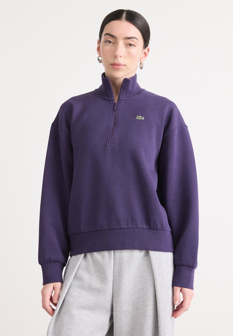 Paarse ritsjack met een hoge kraag en geribbelde boorden, voorzien van een klein groen logo op de linkerborst, gecombineerd met grijze broek.