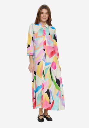 Frau trägt ein farbenfrohes, abstrakt gemustertes, langärmliges Maxikleid mit Knöpfen und schwarze flache Schuhe, steht vor weißem Hintergrund.