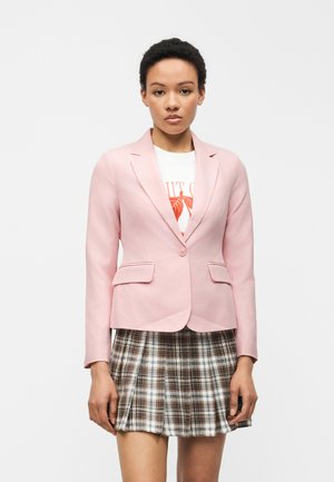 Kvinde iført en lyserød blazer, hvid grafisk t-shirt og brun ternet plisseret nederdel, stående foran en ensfarvet hvid baggrund.