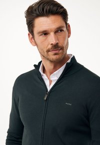Sudadera de cremallera verde oscuro, hecha de un tejido suave, con un cuello alto y un pequeño logo en el pecho. Se lleva sobre una camisa blanca.