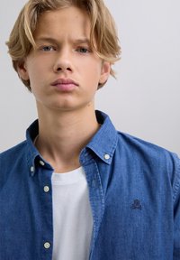 Joven con cabello rubio, vistiendo una camisa blanca y una camisa de mezclilla azul con botones con un pequeño logo de calavera y huesos cruzados.