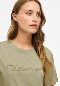 Olijfgroene t-shirt met korte mouwen, een ronde halslijn en een getextureerde "balance" borduring op de borst. Soepele katoenen stof.