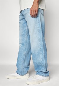 Vaqueros de mezclilla azul claro con corte relajado, que presentan costuras visibles y un corte recto, combinados con zapatillas blancas.
