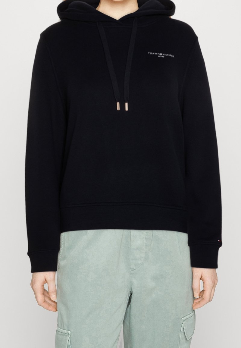 Personne portant un sweat à capuche noir Tommy Hilfiger avec cordons de serrage et un pantalon cargo vert clair, visible du cou jusqu'aux hanches.
