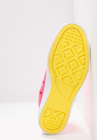 Semelle jaune d'une basket Converse rose inclinée contre un mur blanc, montrant les motifs de la semelle et le marquage en relief sur la semelle en caoutchouc.