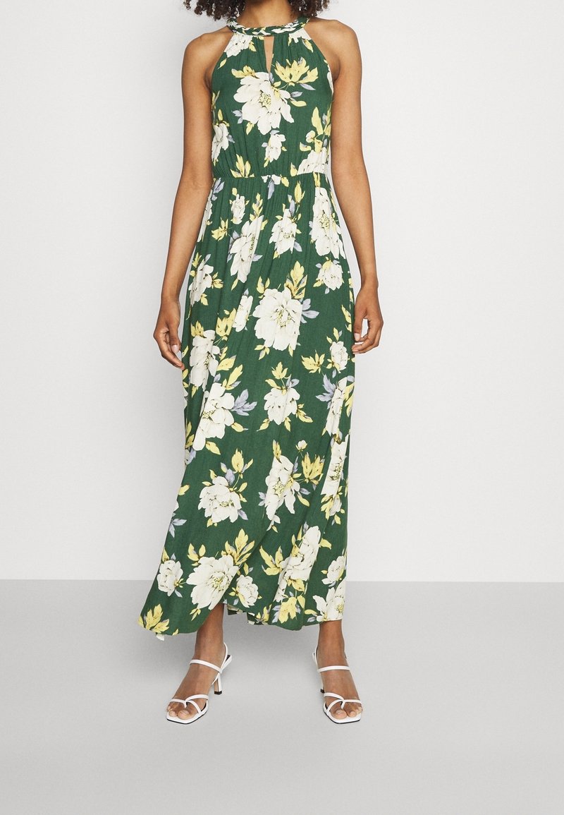 Kvinna i grön ärmlös blommig maxikjol med gula och vita blommor, kombinerad med vita sandalett med klackar.