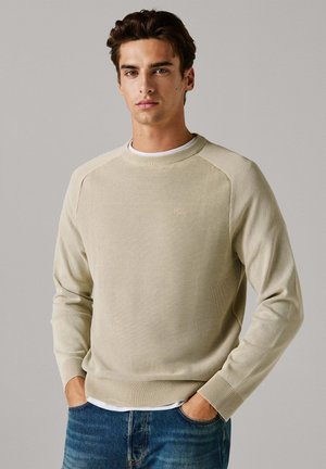 Beež värvusega kootud kampsun soonikkoes mansettide ja äärega, millel on tekstuuriga muster. Kanditud valge T-särgi peal, kombineeritud siniste teksadega.