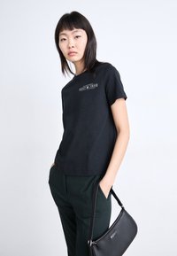 Zwarte katoenen T-shirt met korte mouwen, voorzien van een wit bedrukt logo. Gepaard met donkergroene broeken en een zwarte handtas.