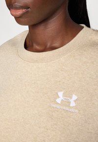 Beige sweatshirt med rund hals, strukturert stoff, og en hvit brodert Under Armour-logo på venstre brystområde.