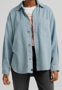 Camicia in corduroy azzurro chiaro con maniche lunghe, chiusura a bottoni sul davanti, taschino sul petto e sottile texture a coste. Indossata sopra una t-shirt grafica.