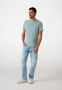 T-shirt de algodón azul claro con cuello redondo y mangas cortas, combinado con jeans de mezclilla de lavado claro y zapatillas blancas. Branding minimal visible.