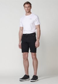 Muž stojí v bielom tričku s krátkym rukávom, čiernych vyhrnutých šortkách a čiernych teniskách, na pozadí svetlo sivej farby.