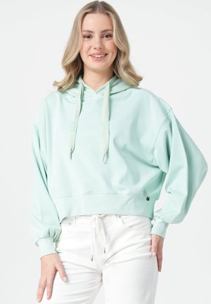Frau mit blondem Haar, die lächelt, trägt einen hellgrünen übergroßen Hoodie und weiße Hosen, steht vor einem schlichten weißen Hintergrund.