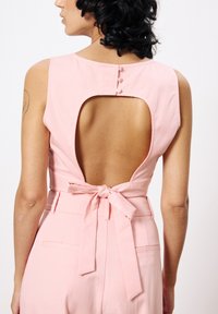 Rosa ärmelloser Jumpsuit mit einem tiefen Rückenausschnitt, einem gebundenen Taillenbereich und Knopfdetails auf der Rückseite. Glatter Stoff und taillierte Passform.