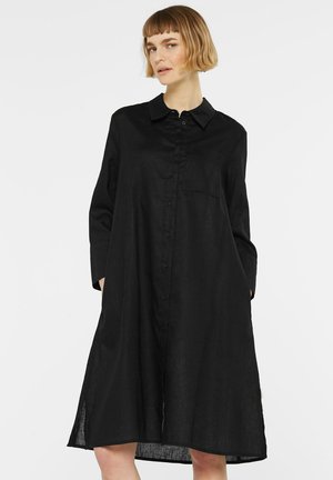 Frau mit kurzen blonden Haaren, die ein langes schwarzes Hemdblusenkleid mit langen Ärmeln trägt und die Hände in den Taschen hat.