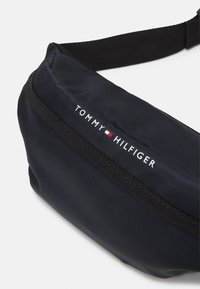 Borsa a tracolla nera in nylon con chiusura a cerniera, caratterizzata dal logo "Tommy Hilfiger" in bianco, insieme ad accenti rossi e blu.