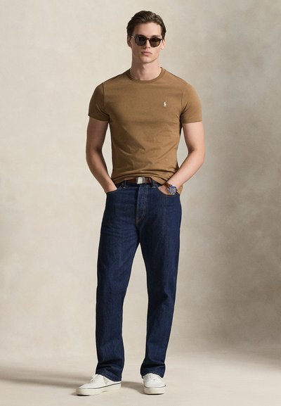 Καφέ εφαρμοστό μπλουζάκι, σκούρο μπλε τζιν και άσπρα αθλητικά παπούτσια. Το μοντέλο φορά γυαλιά ηλίου και ρολόι χειρός, στέκεται μπροστά σε ουδέτερο φόντο.