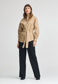 Beige Hemd mit Knopfverschluss, einem taillierten Bund, langen Ärmeln und Manschettendetails, kombiniert mit schwarzen weit geschnittenen Hosen. Glatter Stoff und taillierte Passform.