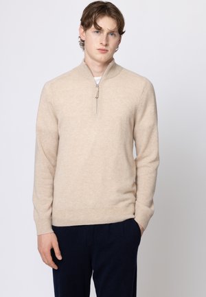 Giovane uomo con capelli castano chiaro che indossa un maglione beige con cerniera e pantaloni scuri, in piedi contro uno sfondo chiaro e semplice.