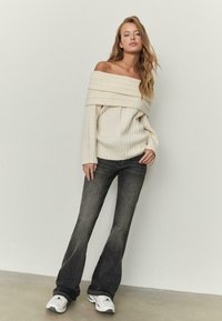 Krem pulover z rebrastim vzorcem in odprtimi rameni, z dolgimi rokavi, kombiniran s temnimi razširjenimi kavbojkami. Model nosi bele superge in stoji na nevtralnem ozadju.