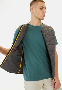 Grå polstret vest med kviltet design, der har en gul lynlåsinventar. Båret over en teal t-shirt og vist mod en ensfarvet baggrund.