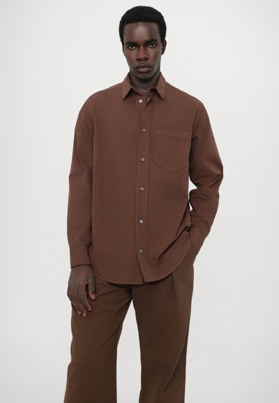 Chemise brune à boutons avec une poche sur la poitrine et des manches longues, en tissu doux. Associée à un pantalon large assorti de couleur brune.
