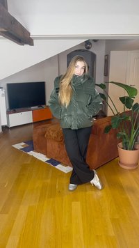 Mlada žena u zelenoj jakni na preklop, crnim hlačama i srebrnim tenisicama stoji prekriženih nogu u dnevnom boravku koji ima sofu, televizor i veliku biljku.