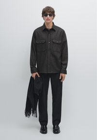 Giacca camicia in lana scura con due tasche sul petto, indossata sopra una maglia chiara, abbinata a pantaloni e scarpe neri, mentre tiene una sciarpa nera.