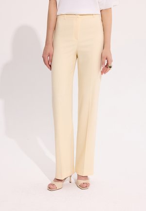 Kobieta nosząca kremowe spodnie z wysokim stanem i prostą nogawką, biały top i beżowe sandały na obcasie z odkrytymi palcami, stojąca na jednolitym tle.