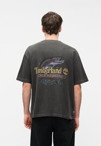 Άντρας που φοράει σκούρα γκρι φαρδιά μπλουζά T-shirt με πολύχρωμο γραφικό ψαριού και το κείμενο «Timberland Catch and Release!» στην πλάτη.