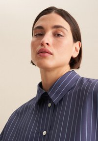 Camicia blu navy a maniche corte con righe verticali azzurre, colletto a punta e due bottoni, caratterizzata da una texture di tessuto liscia.