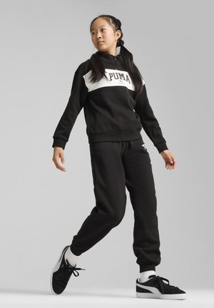 Felpa nera con pannello bianco e logo sul petto; abbinata a pantaloni jogger neri coordinati. Sneaker nere con dettagli bianchi e chiusura con lacci.