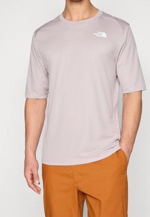 Homme portant un t-shirt North Face gris clair à manches courtes et un pantalon orange, une main dans la poche, fond neutre.