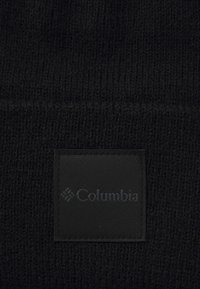 Black knitted beanie with a ribbed texture, featuring a rectangular black patch with "Columbia" logo. Soft and durable material.