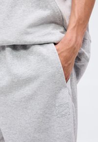 Gros plan de la main d’une personne dans la poche d’un pantalon de survêtement gris clair avec un logo Nike Athletic Club en relief subtil.